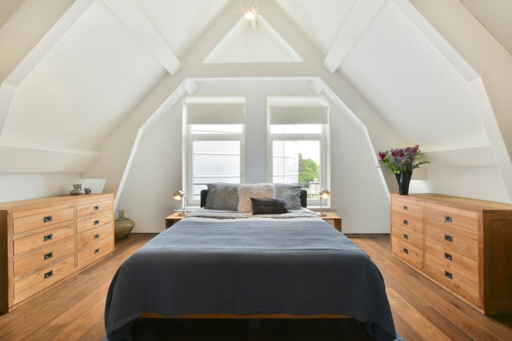 A modern, remodeled attic bedroom with angled ceilings, twin windows, and wooden furniture—showcasing Reflex Builders' attic remodeling services.