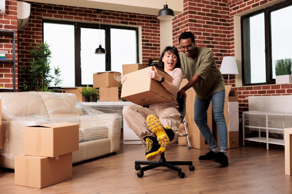 A joyful couple surrounded by moving boxes, with one person pushing the other on an office chair—illustrating the excitement and chaos of deciding between moving or renovating.