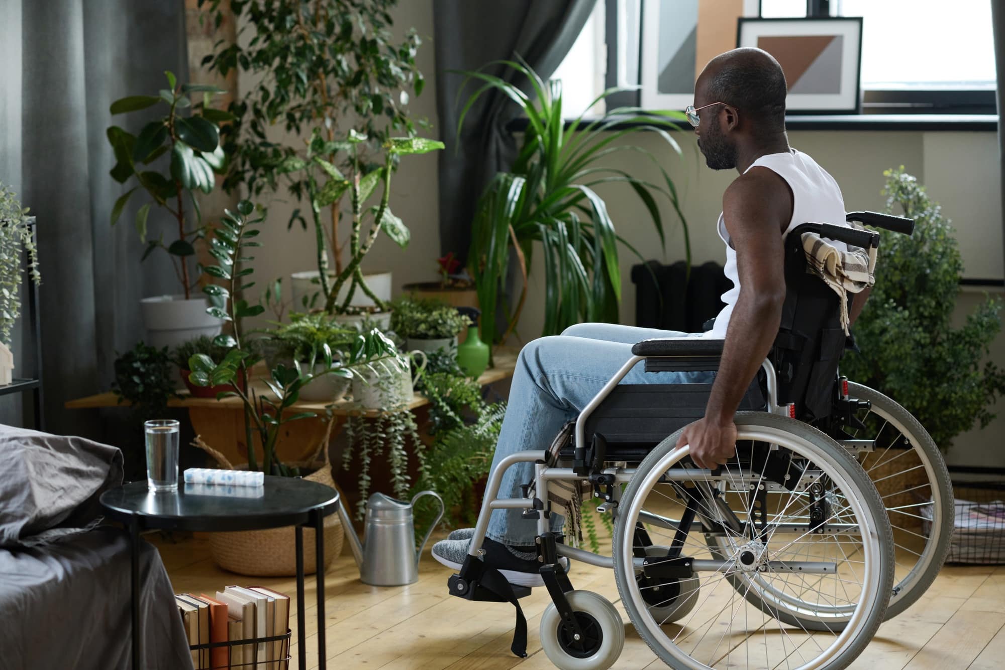 Man in a wheelchair inside a bright, plant-filled room, showcasing elements of accessible home design with wide open spaces and reachable surfaces.