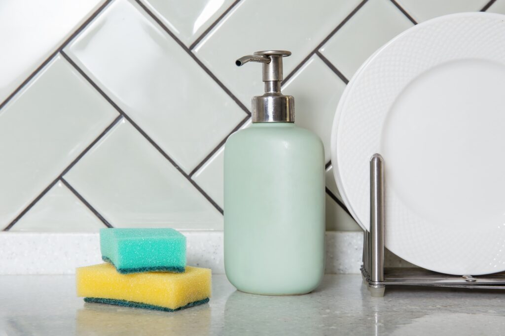 Close-up of a glossy herringbone tile pattern behind a soap dispenser, sponge, and dish rack&mdash;illustrating unique kitchen backsplash ideas.