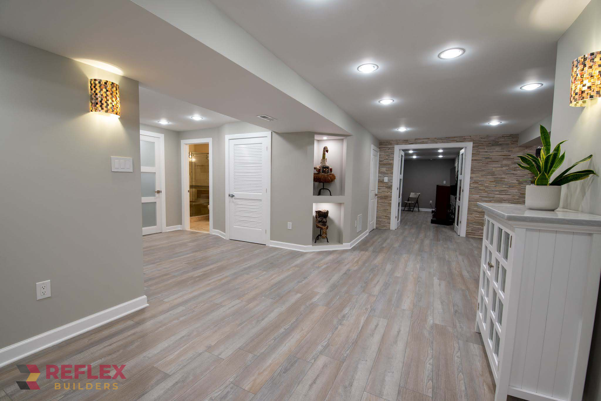 A beautifully finished modern basement remodel by Reflex Builders, featuring light gray walls, gray wood-look vinyl flooring, built-in display niches, recessed LED lighting, stone accent walls, and decorative wall sconces.