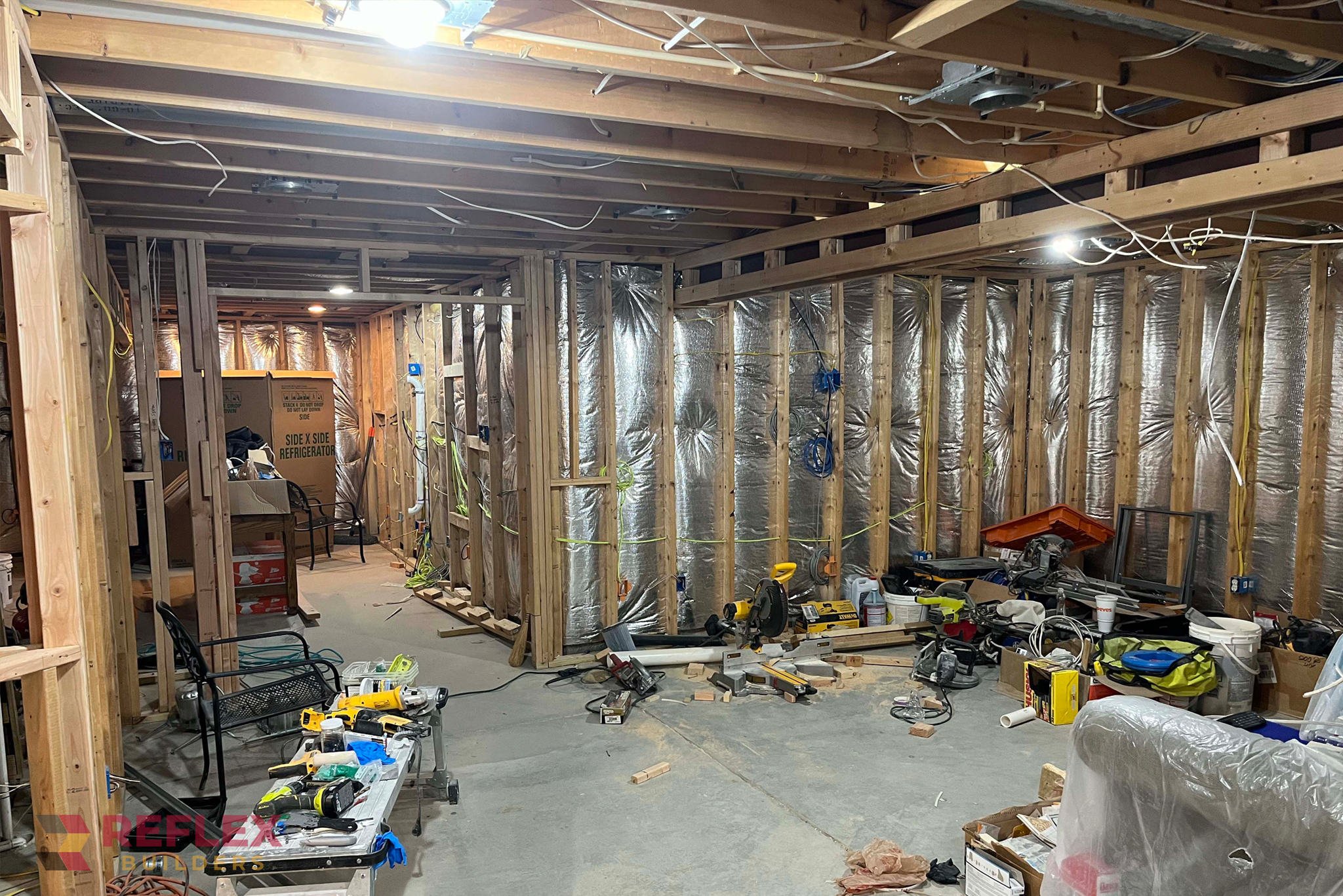 An unfinished basement undergoing a full renovation by Reflex Builders, featuring exposed wood framing, reflective wall insulation, rough-in electrical wiring, and various construction tools across a concrete floor.